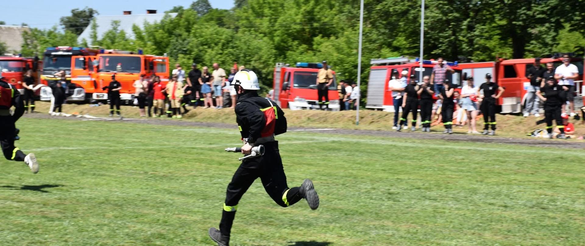 Zdjęcie przedstawia biegnącego po trawie strażaka. Strażak ubrany w czarny mundur i biały hełm, w lewej ręce trzyma prądownicę. W tle niewyraźnie widać widzów oraz czerwone samochody pożarnicze.