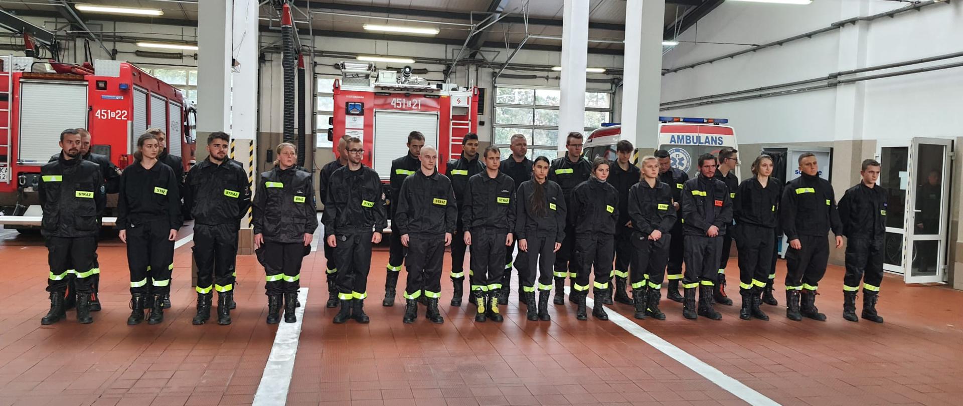 Dwuszereg strażaków ratowników jednostek Ochotniczej Straży Pożarnej oczekujący na wręczenie dyplomów. Scena rozgrywa się na terenie garażów Jednostki Ratowniczo Gaśniczej w Kamiennej Górze w tle ustawione samochody ratowniczo gaśnicze.