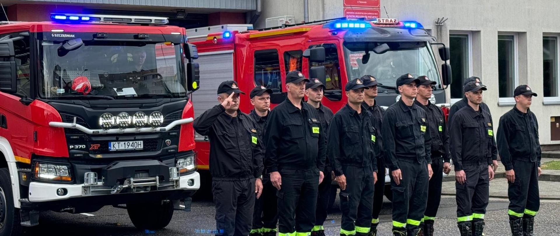 Strażacy stoją ustawienie w dwuszeregu, ubrani w czarne ubrania koszarowe, na głowach mają czapeczki czarne z daszkiem. Za strażakami ustawione są czerwone samochody pożarnicze, na tle garaży.