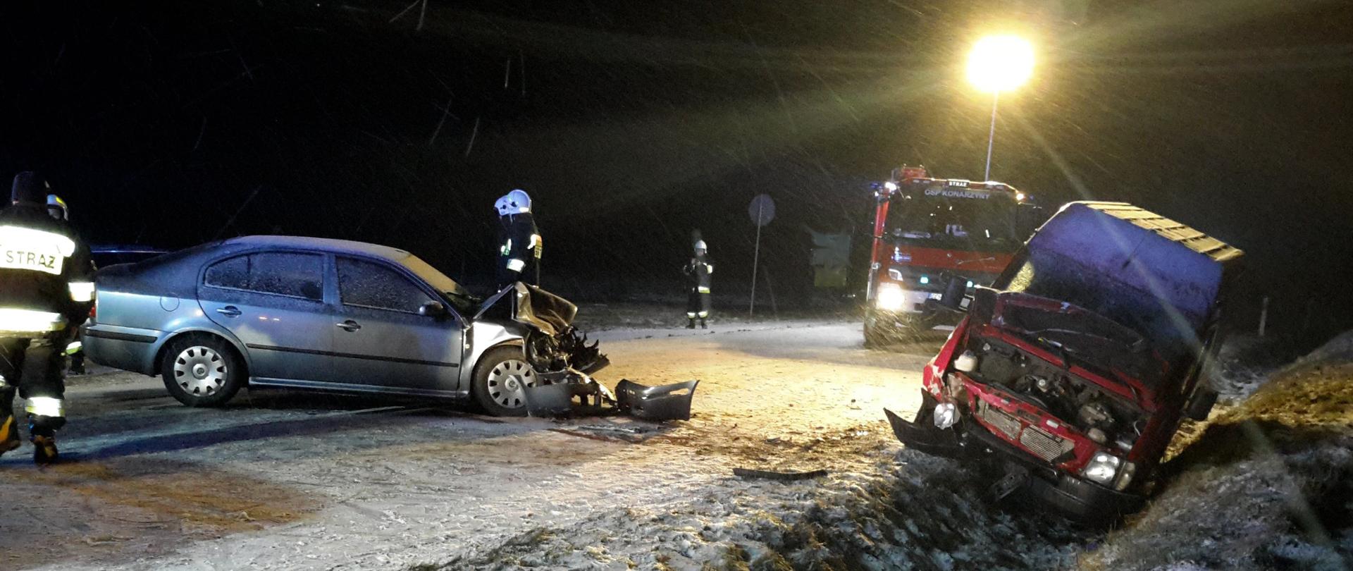 Czerwony samochód dostawczy w otwartą pokrywą silnika leży w rowie na poboczu drogi. Pogięte zderzak oraz uszkodzone lampy. Obok, na drodze znajduje się samochód osobowy z rozbitym przodem. Wokół strażacy uczestniczący w działaniach ratowniczych. W tle widać wóz strażacki oświetlający teren zdarzenia.