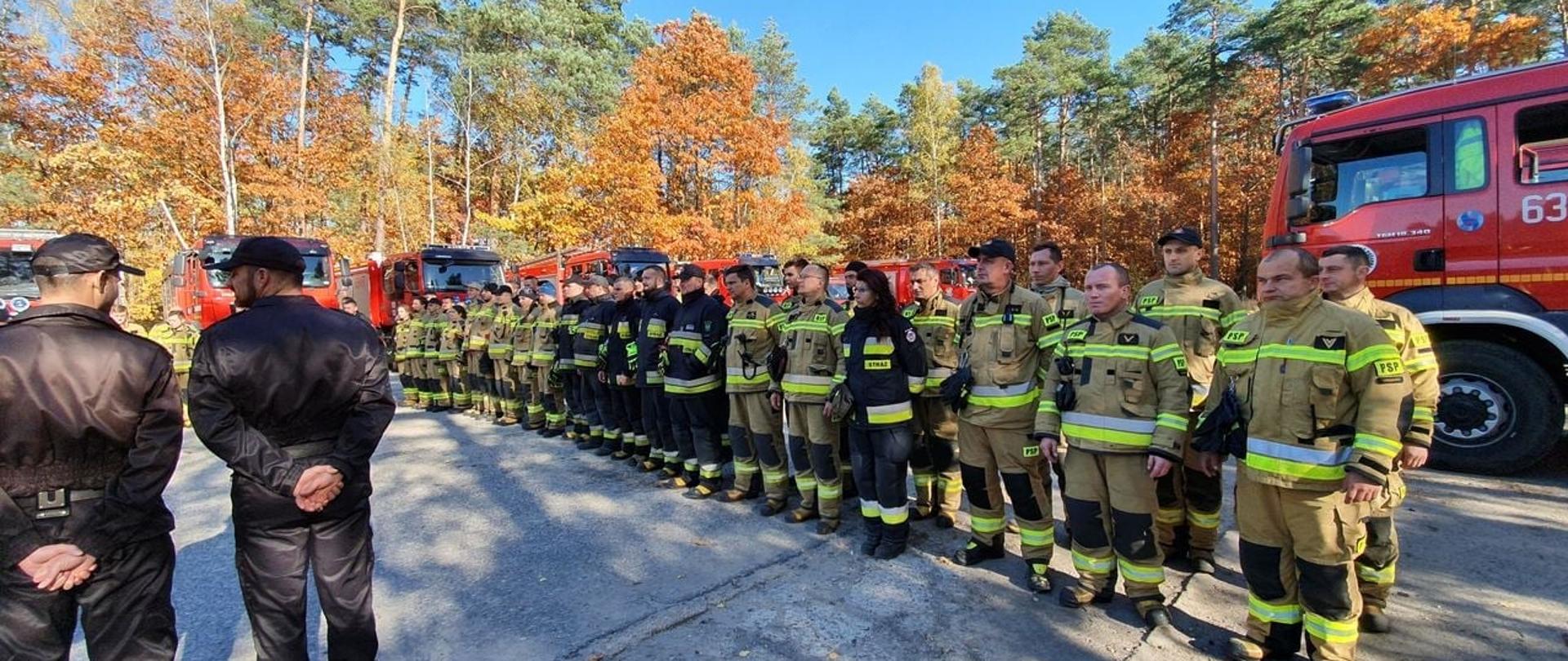 Po lewej dwóch strażaków w ubraniu dowódczo sztabowym , po prawej pododdział strażaków w dwuszeregu, Z tyłu kilka samochodów ratowniczo-gaśniczych na tle drzew
