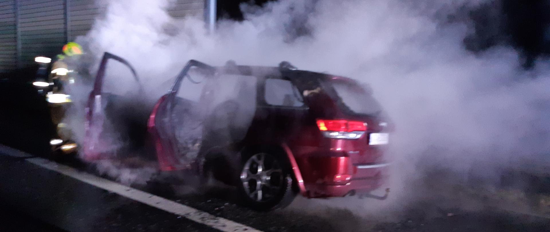 Na zdjęciu samochód osobowy, który się pali. Strażacy w aparatach ochrony układu oddechowego prowadzą działania gaśnicze