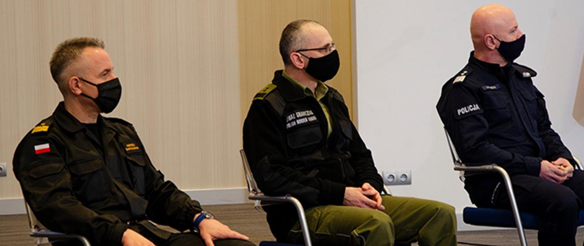 Trzy osoby w mundurach siedzą na krzesłach w maseczkach na twarzach.