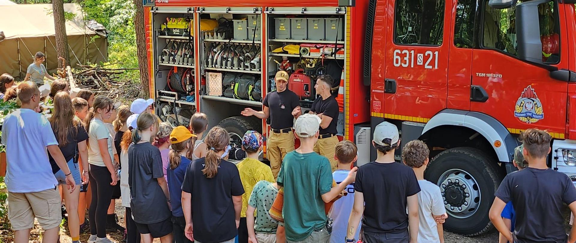 Grupa dzieci stoi przodem w kierunku samochodu strażackiego czerwonego z otwartymi skrytkami.