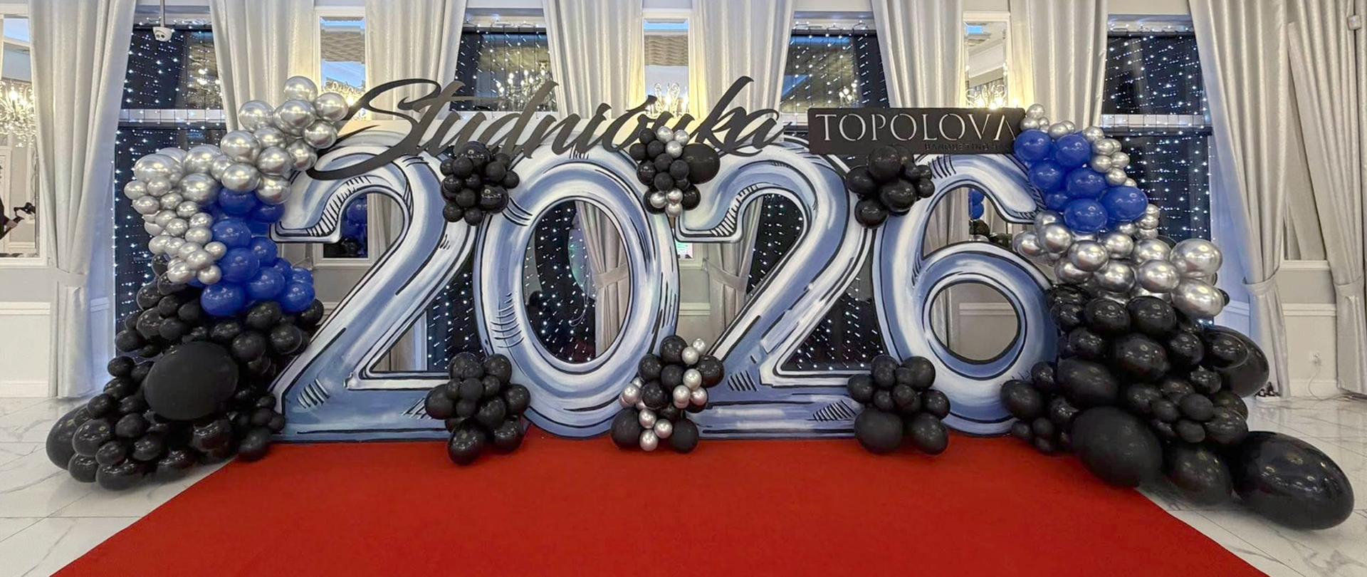 Ściankę fotograficzną na tle okien, z balonów i dużym napisem 2026, nad cyframi napis Studniówka i Topolova.