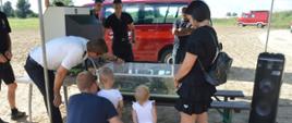 Piknik "Kręci mnie bezpieczeństwo nad wodą". Plaża na byłym poligonie wojskowym w Sierakowie. Namiot, pod którym strażacy z KP PSP w Rawiczu edukują uczestników imprezy w zakresie przestrzegania zasad bezpieczeństwa podczas wypoczynku nad wodą i w lesie. Na pierwszym planie mobilny symulator zagrożeń w postaci modułu woda-las. W zajęciach edukacyjnych uczestniczą zarówno dzieci jak i osoby dorosłe.W tle drzewa oraz samochody pożarnicze