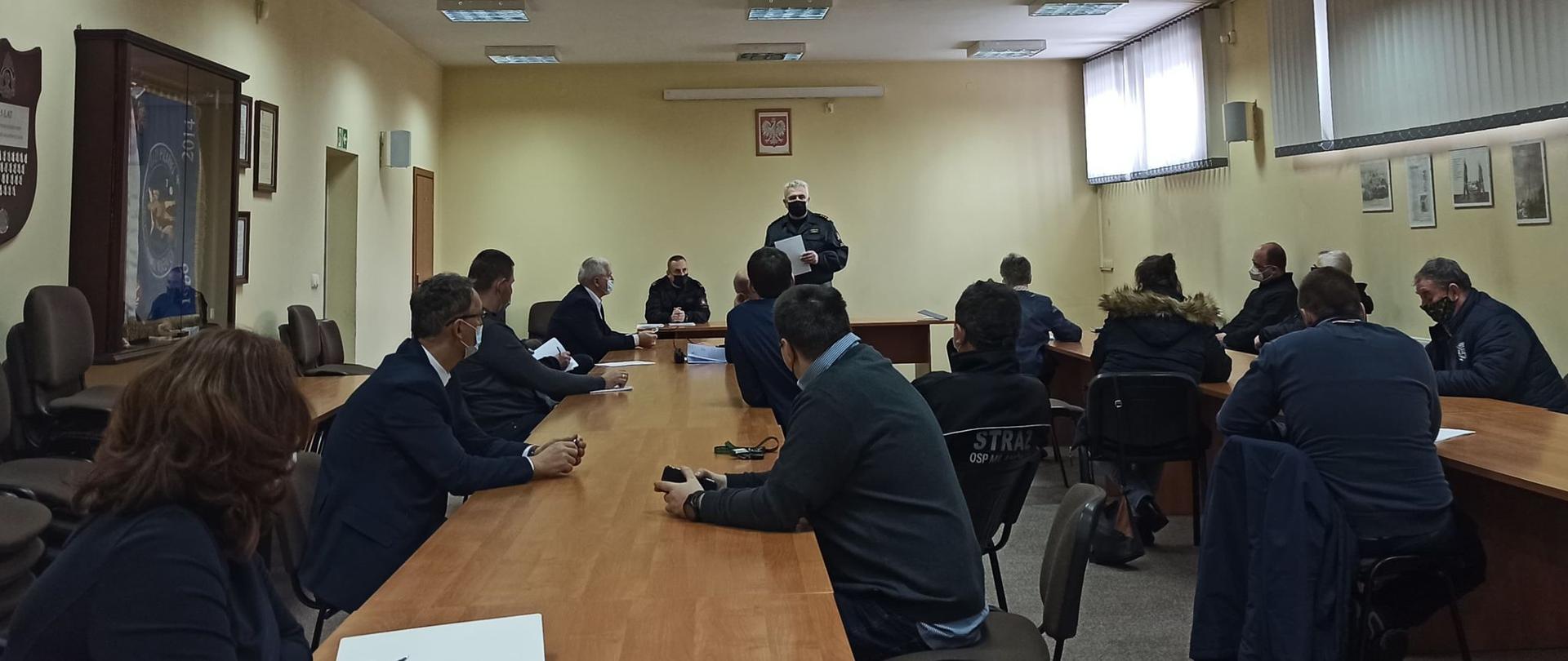 Przedstawiciele samorządu lokalnego oraz Ochotniczych Straży Pożarnych siedząc na krzesłach przy stołach na sali konferencyjnej słuchają wystąpienia Komendanta Powiatowego Państwowej Straży Pożarnej w Grodzisku Mazowieckim dotyczącego koordynacji wspólnych działań w zakresie wsparcia dla seniorów w dotarciu do punktów szczepień na COVID- 19 oraz przeprowadzenia dystrybucji materiałów informacyjnych dotyczących Narodowego Programu Szczepień przeciw Covid-19 wśród seniorów z rejonu powiatu grodziskiego.