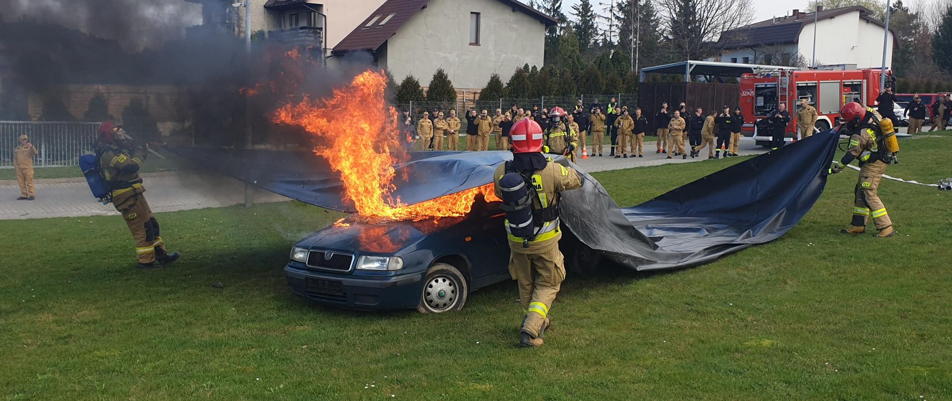 Strażacy w ubrani w nomexy i aparaty ochrony dróg oddechowych naciągają na palący się samochód płachtę gaśniczą. W tle znajduje się samochód ratowniczo - gaśniczy z rozwiniętą linią gaśniczą oraz strażacy obserwujący pokaz.