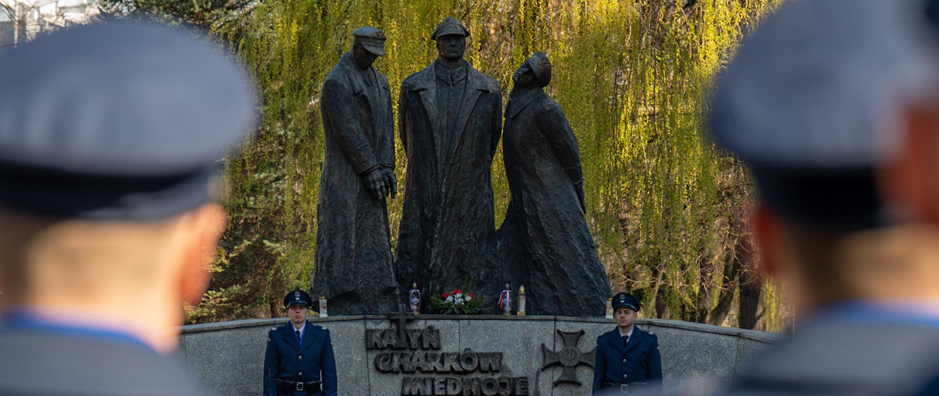 Zdjęcie przedstawia pomnik znajdujący się na otwartej przestrzeni, w parku, o czym świadczą widoczne w tle drzewa. Na pierwszym planie, w dolnej części kadru, widzimy dwie osoby w mundurach, stojące tyłem do obserwatora. Są to policjanci w mundurach. Mundury mają ciemnoszary kolor, a na głowach widoczne są czapki z daszkiem. Osoby te stoją symetrycznie, po obu stronach centralnej osi zdjęcia, tworząc swego rodzaju ramę dla dalszej części obrazu.
W centrum kadru, na drugim planie, znajduje się pomnik ofiar katynia. Składa się on z trzech rzeźbionych postaci, które wydają się być ludźmi. Postacie te są ubrane w długie płaszcze, a ich sylwetki są smukłe i wyciągnięte. Rzeźby mają ciemny, szary kolor, co nadaje im melancholijny i poważny wygląd. Bezpośrednio pod rzeźbami znajduje się cokół, na którym umieszczono tablicę z napisem. Niestety, treść napisu jest nieczytelna z powodu odległości i perspektywy.