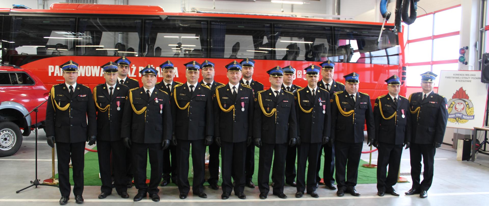 Zdjęcie przedstawia sylwetki strażaków odchodzących na zaopatrzenie emerytalne w asyście komendanta miejskiego Państwowej Straży Pożarnej oraz jego zastępcy, ubranych w mundury wyjściowe w postawie zasadniczej. Strażacy stoją w dwóch szeregach na tle wozu - autobusu strażackiego w garażu Jednostki Ratowniczo-Gaśniczej Nr 3 w Rzeszowie.