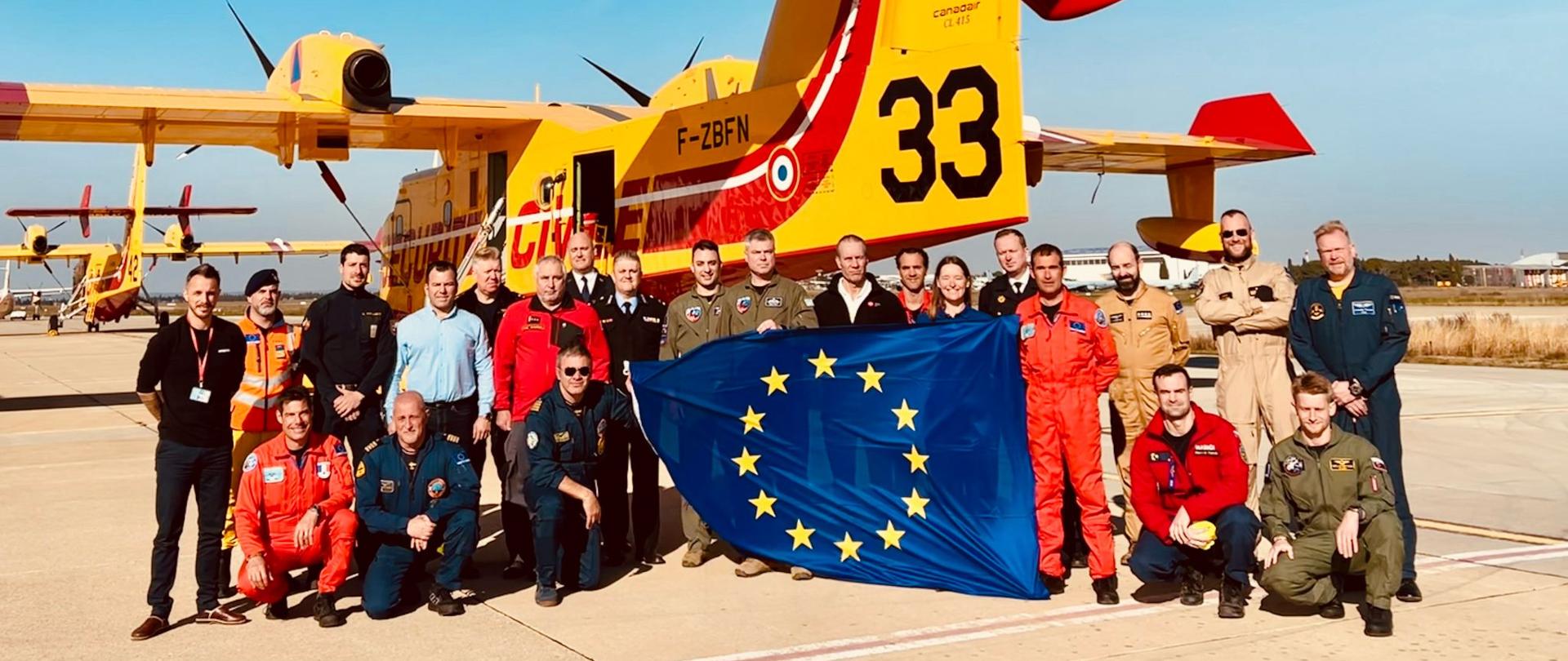 Zdjęcie pamiątkowe uczestników spotkania na tle samolotu Canadair. Pozujące do zdjęcia osoby są w różnego rodzaju umundurowaniu a część z nich trzyma flagę Unii Europejskiej