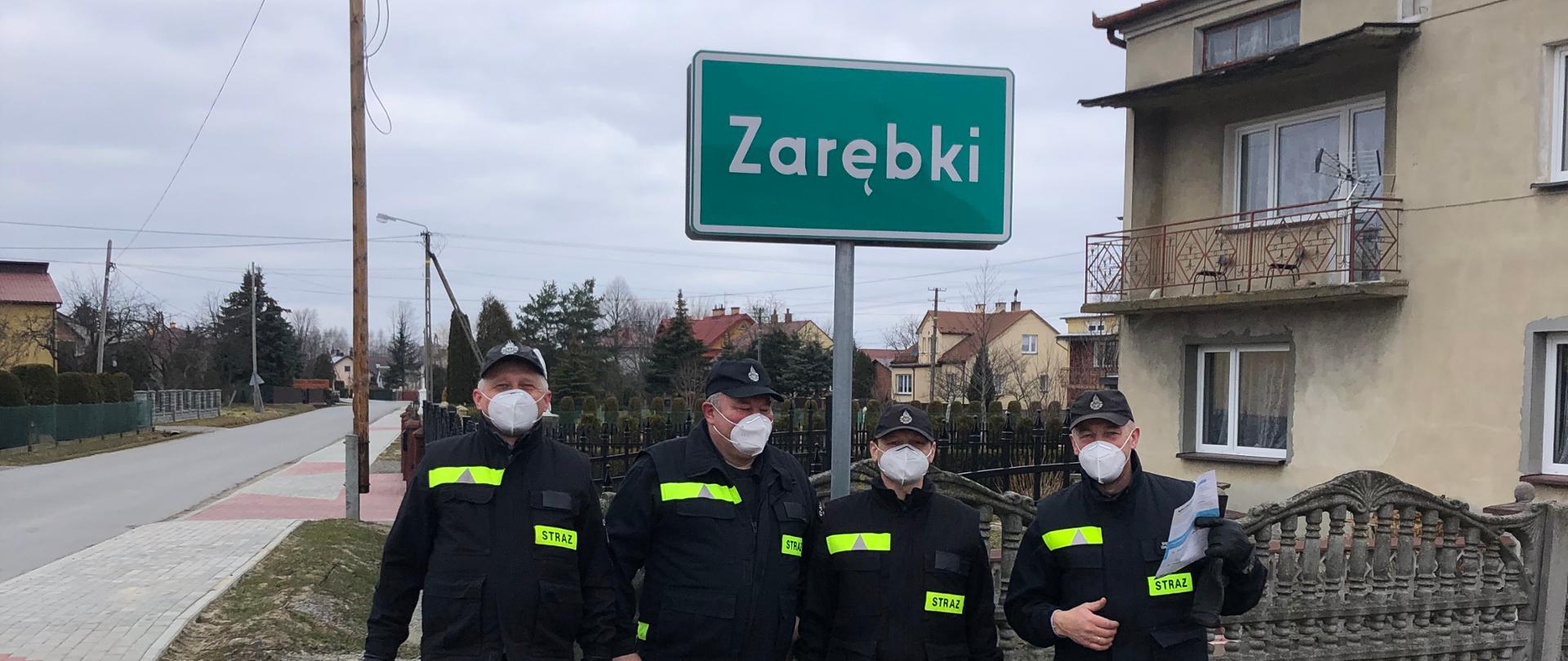 Na zdjęciu czterech strażaków stoi przy znaku miejscowości Zrębki. W tle po lewej stronie zdjęcia fragment chodnika i drogi asfaltowej, po prawej fragment płotu i budynek