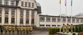 Strażacy ustawieni w dwuszeregu po prawej stronie maszty flagowe przed którymi stoi poczet flagowy
