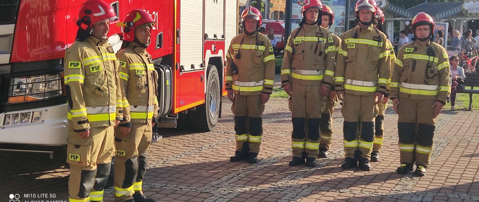 Strażacy w żółtych mundurach bojowych stoją w dwuszeregu na tle samochodu pożarniczego. za nimi w prawej części zdjęcia ludzie chodzący po rynku.