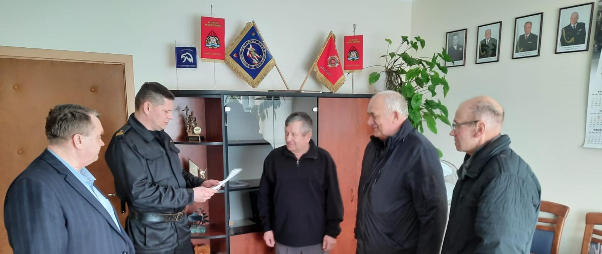 Zdjęcie przedstawia Komendanta Powiatowego Państwowej Straży Pożarnej w Zambrowie w ubraniu dowódczo-sztabowym wręczającego trzem druhom w ubraniach cywilnych decyzję o przyznaniu świadczenia ratowniczego. Z prawej strony stoi pracownik Urzędu Miasta Pan Bogdan Pac. Zdjęcie wykonano w gabinecie Komendanta Powiatowego. 