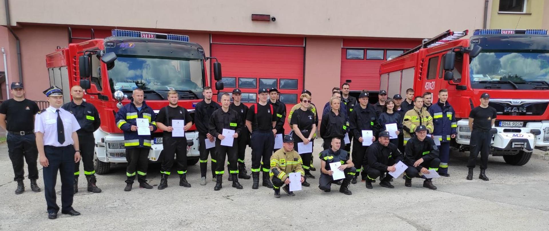Przed budynkiem strażnicy na tle dwóch czerwonych wozów bojowych stoją strażacy trzymając w ręku zaświadczenia ukończenia kursu.