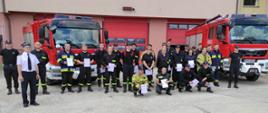 Przed budynkiem strażnicy na tle dwóch czerwonych wozów bojowych stoją strażacy trzymając w ręku zaświadczenia ukończenia kursu.