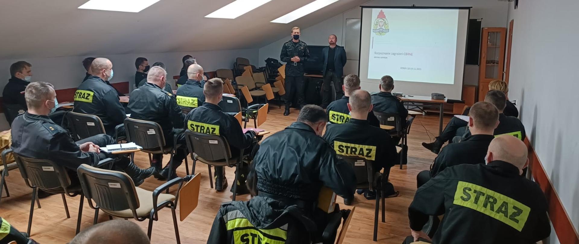 grupa strażaków w mundurach siedzi w sali, na przeciwko stoi dwóch meżczyzn, w tle prezentacja, wszyscy w maseczkach na twarzach