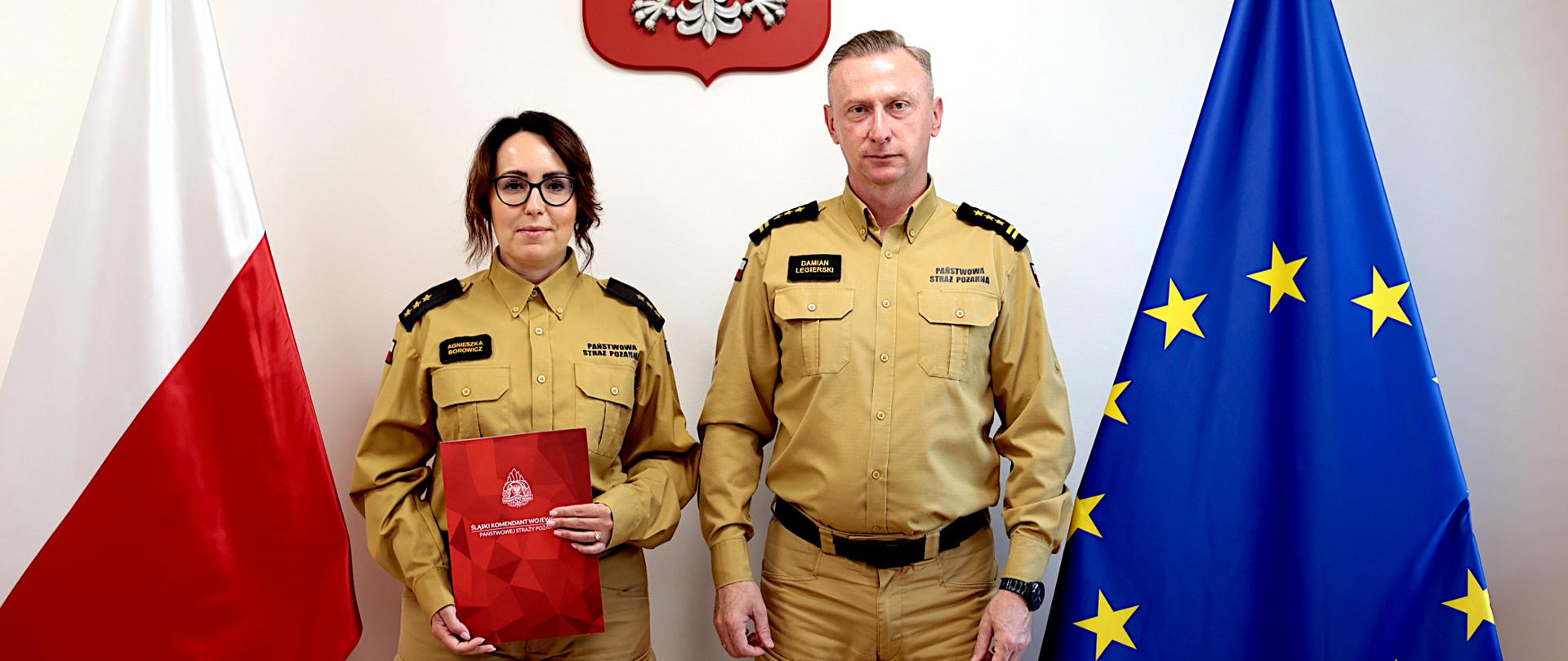 Na zdjęciu stoją obok siebie w piaskowych mundurach służbowych - Główna Księgowa kpt. Agnieszka Borowicz w Komendzie Wojewódzkiej i śląski komendant wojewódzki st. bryg. Damian Legierski. Kobieta trzyma czerwoną teczkę z dokumentem mianowania na stanowisko głównej księgowej. W tle widać polską flagę po lewej stronie, flagę Unii Europejskiej po prawej oraz godło Polski zawieszone na ścianie nad nimi.