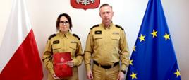 Na zdjęciu stoją obok siebie w piaskowych mundurach służbowych - Główna Księgowa kpt. Agnieszka Borowicz w Komendzie Wojewódzkiej i śląski komendant wojewódzki st. bryg. Damian Legierski. Kobieta trzyma czerwoną teczkę z dokumentem mianowania na stanowisko głównej księgowej. W tle widać polską flagę po lewej stronie, flagę Unii Europejskiej po prawej oraz godło Polski zawieszone na ścianie nad nimi.