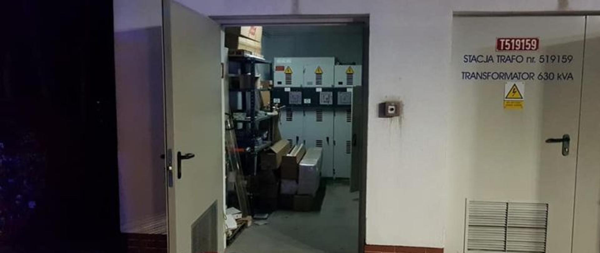 Zdjęcie przedstawia pomieszczenie w którym wybuchł pożar od zewnątrz.Pomieszczenie ma otwarte drzwi. W środku pali się światło przy drzwiach stoi gaśnica. Zdjęcie wykonane po zmierzchu.
