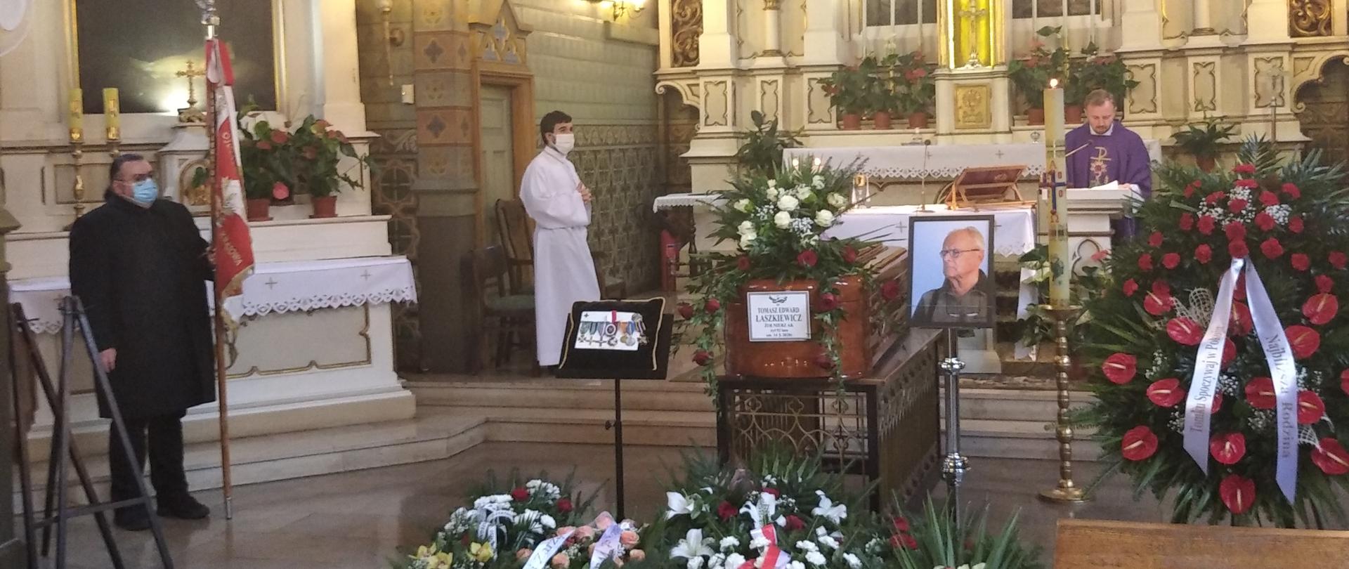 Zdjęcie zrobione jest w kościele podczas pogrzebu. Znajduje się na nim trumna oraz wiązanki kwiatów.