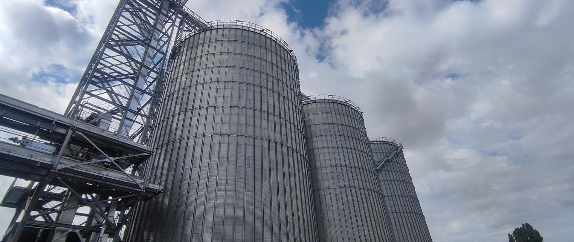 Na zdjęciu widać trzy żelbetowe silosy służące do przechowywania zboża. Na drugim planie pochmurne niebo.