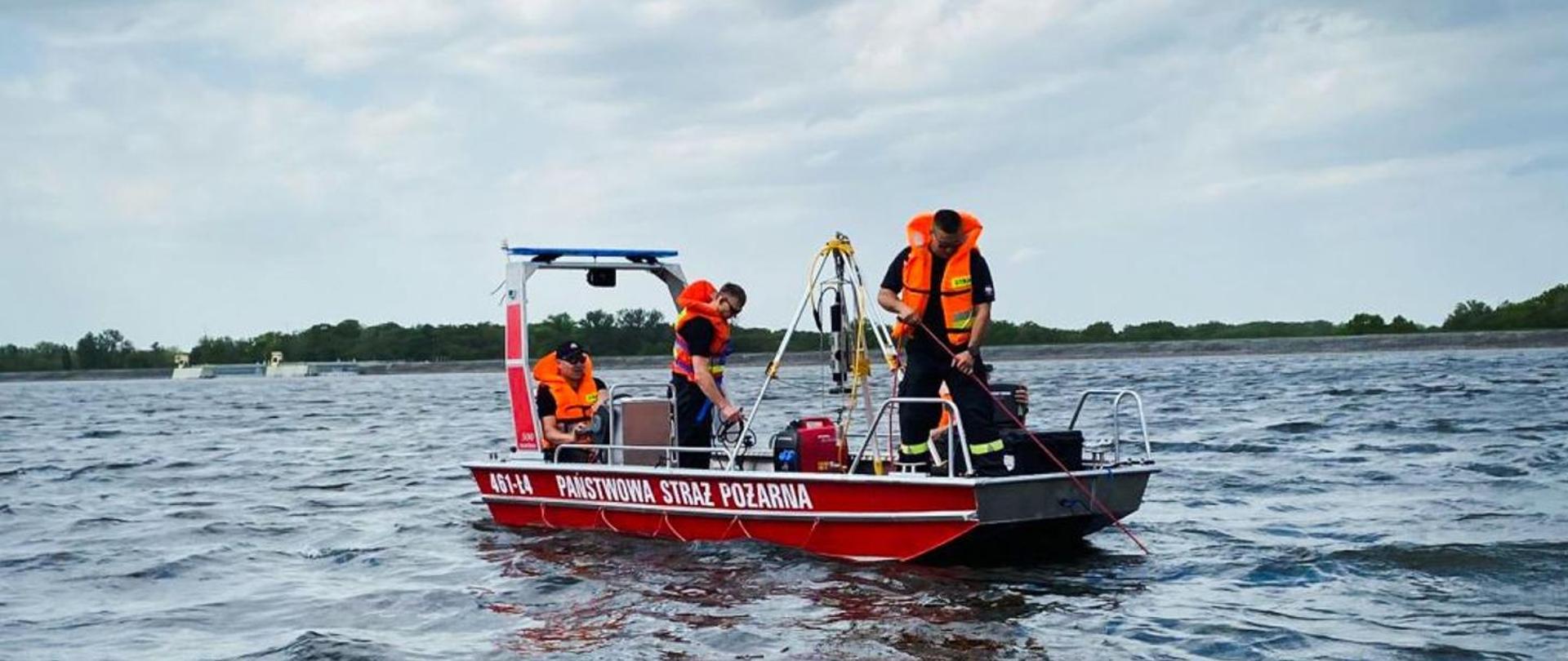 Trzech strażaków w łódce na jeziorze. Dwóch stoi, jeden siedzi