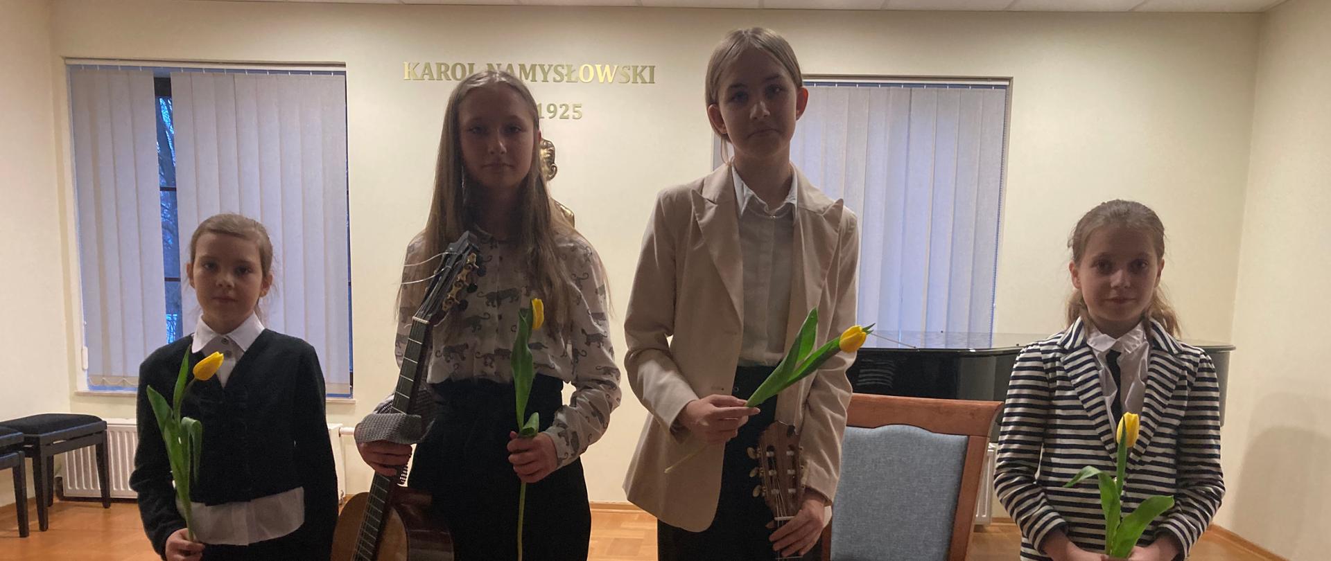 Uczennice klasy gitary pani Iwony Grabowskiej po "Koncercie gitarowym" w dniu 8 masrca 2023 r. – od lewej strony Lena Torba, Wiktoria Szymona, Maria Gimel, Daria Samulak stoją na scenie Sali koncertowej szkoły muzycznej. Dziewczynki ubrane są na galowo, trzymają w ręku żółte tulipany, które dostały za występ. Dwie dziewczynki w środku trzymają w ręku gitary. 