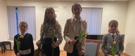 Uczennice klasy gitary pani Iwony Grabowskiej po "Koncercie gitarowym" w dniu 8 masrca 2023 r. – od lewej strony Lena Torba, Wiktoria Szymona, Maria Gimel, Daria Samulak stoją na scenie Sali koncertowej szkoły muzycznej. Dziewczynki ubrane są na galowo, trzymają w ręku żółte tulipany, które dostały za występ. Dwie dziewczynki w środku trzymają w ręku gitary. 
