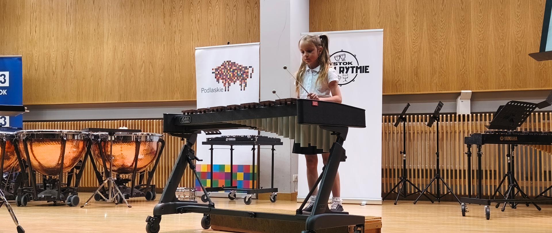 Zdjęcie przedstawia scenę sali koncertowej Państwowej Szkoły Muzycznej I i II stopnia w Białymstoku.
Na zdjęciu widzimy grającą na ksylofonie Adriannę Graszę podczas I Ogólnopolskiego Konkursu Perkusyjnego "Białystok w Rytmie". Z lewej strony sceny widzimy kotły orkiestrowe. Centralnie z tyłu sceny stoją banery I Ogólnopolskiego Konkursu Perkusyjnego "Białystok w Rytmie". Z prawej strony sceny widzimy ksylofon i trzy pulpity.

