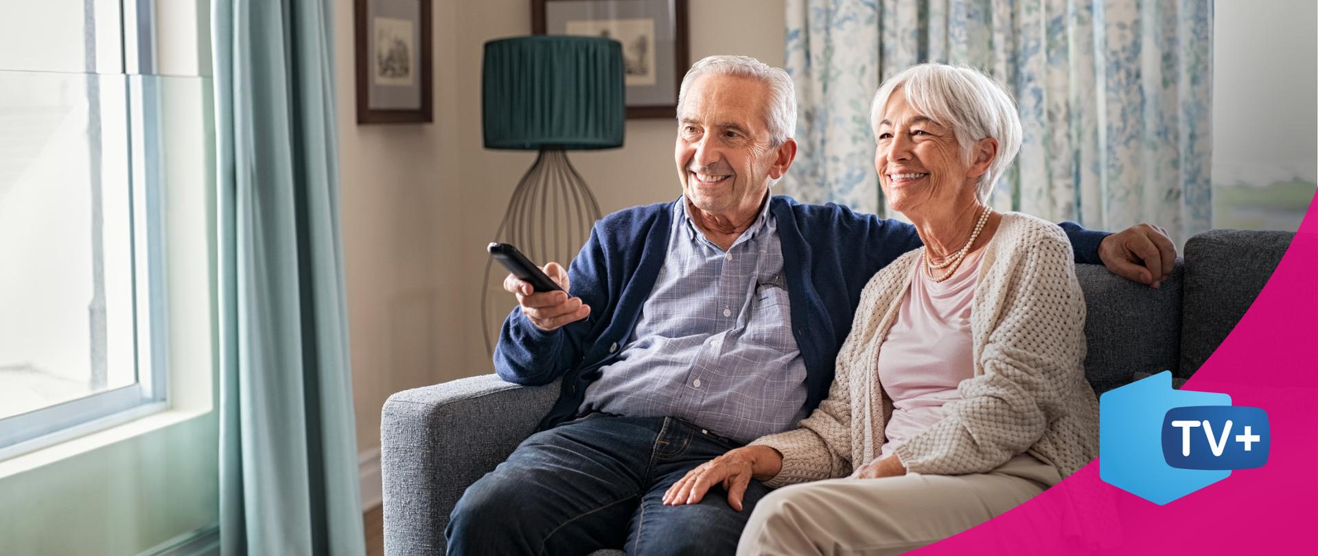 zdjęcie przedstawiające parę starszych ludzi siedzących na kanapie, uśmiechają się. Mężczyzna trzyma pilota do telewizora w ręku.