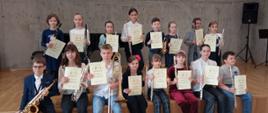 Grupa uczniów siedzi i stoi na scenie sali koncertowej, w rękach trzymają instrumenty i dyplomy