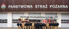 Na zdjęciu widać grupę strażaków stojących przed budynkiem z dużym napisem „Państwowa Straż Pożarna”. Przed budynkiem zaparkowany wóz strażacki drabiną mechaniczną. Strażacy ustawili się do wspólnego zdjęcia grupowego 