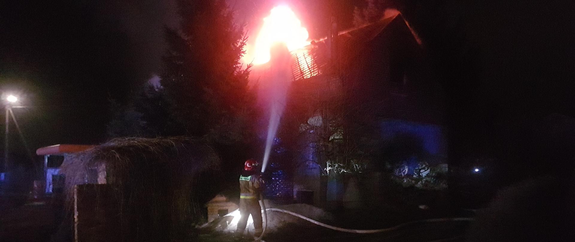 Zdjęcie przedstawia strażaka podającego prąd wody na palący się budynek. 