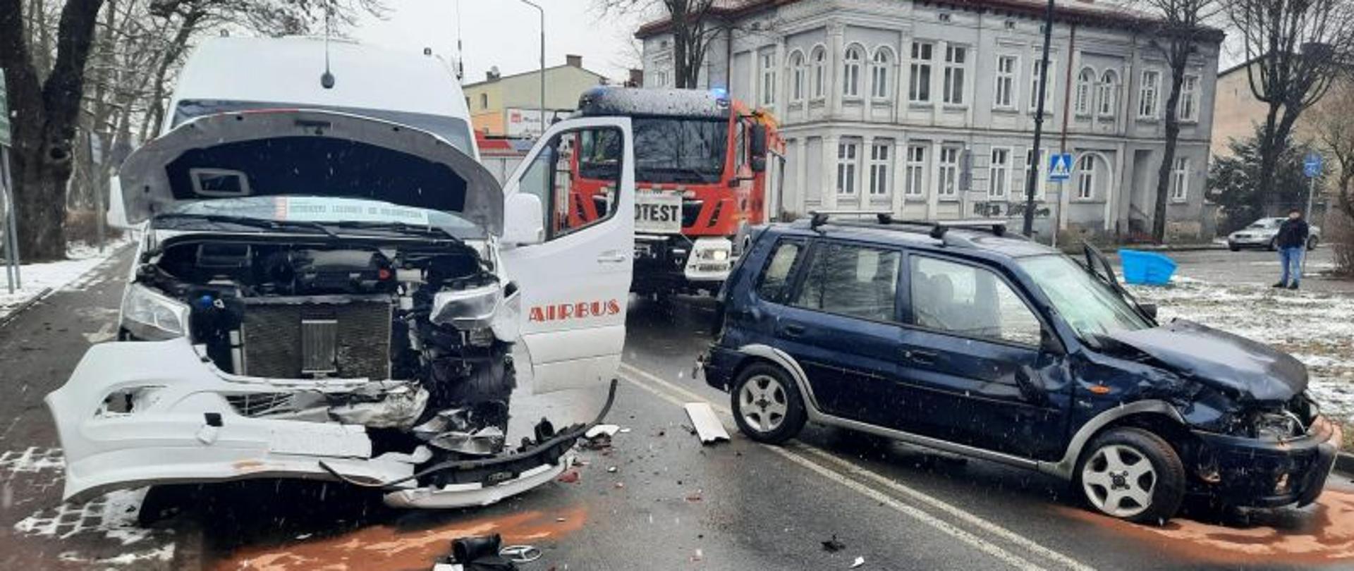 Widok na miejsce zdarzenia. Centralny punkt zdjęcia stanowią trzy auta.Dwa uczestniczące w zdarzeniu, biały bus oraz terenówka. W tle wóz strażacki. Jest mroźno. Auta mają rozbite przody.