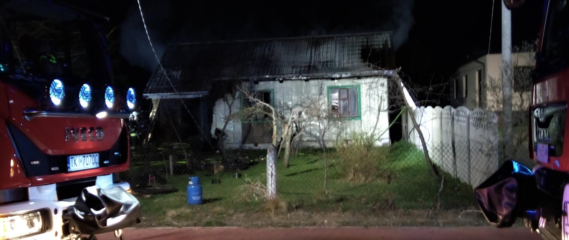 Fotografia przedstawia jednopiętrowy, drewniany budynek mieszkalny, w którym wybuchł pożar na drugim planie. Na pierwszym planie po lewej i prawej stronie domu stoją samochody pożarnicze.