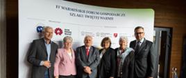 Grupa oficjeli pozuje do wspólnego zdjęcia. W centrum stoi wicewojewoda w szarym garniturze i czerwonym krawacie. W tle widoczny napis „Warmińskie Forum Gospodarcze Szlaku Świętej Warmii”.