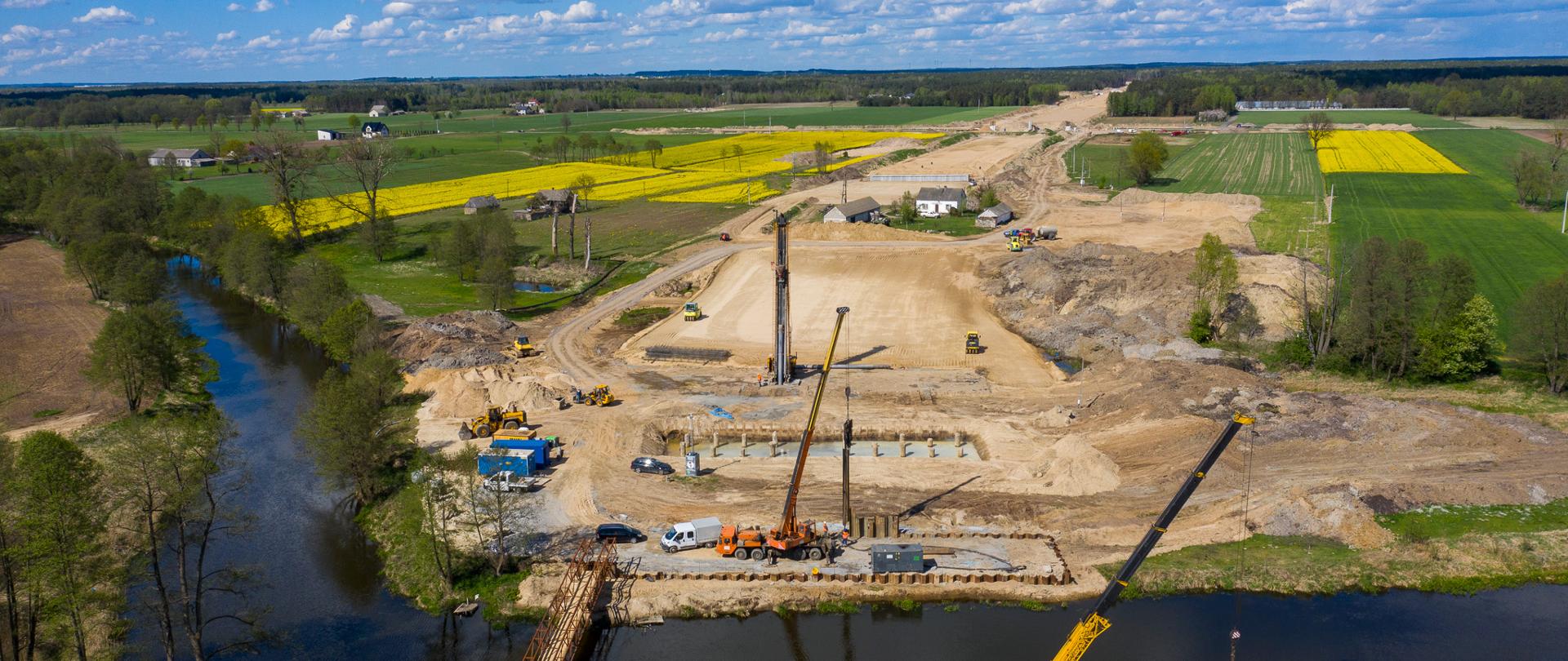 Lotnicze ujęcie placu budowy drogi wraz z powstającym mostem nad rzeką; widoczne dźwigi, maszyny budowlane oraz rozległe pola i zabudowania w tle.