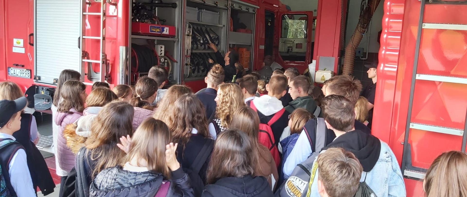 3. Zdjęcie numer trzy przedstawiam jedną wielką grupę uczniów przy dwóch strażakach. Uczniowie oraz nauczycielki chętnie oglądają sprzęty na samochodach oraz jest on omawiany. Jeden strażak w chwili robienia zdjęcia wyjmuje wąż strażacki w celu jego zaprezentowania oraz omówienia drugi zaś strażak chętnie odpowiada na pozostałe pytanie zadawane przez młodzież. Wszyscy znajdują się w garażu podziału bojowego jednostki ratowniczo-gaśniczej w Aleksandrowie kujawskim i stoją między dwoma wozami strażackimi z otwartymi roletami na sprzęty ratownicze. 