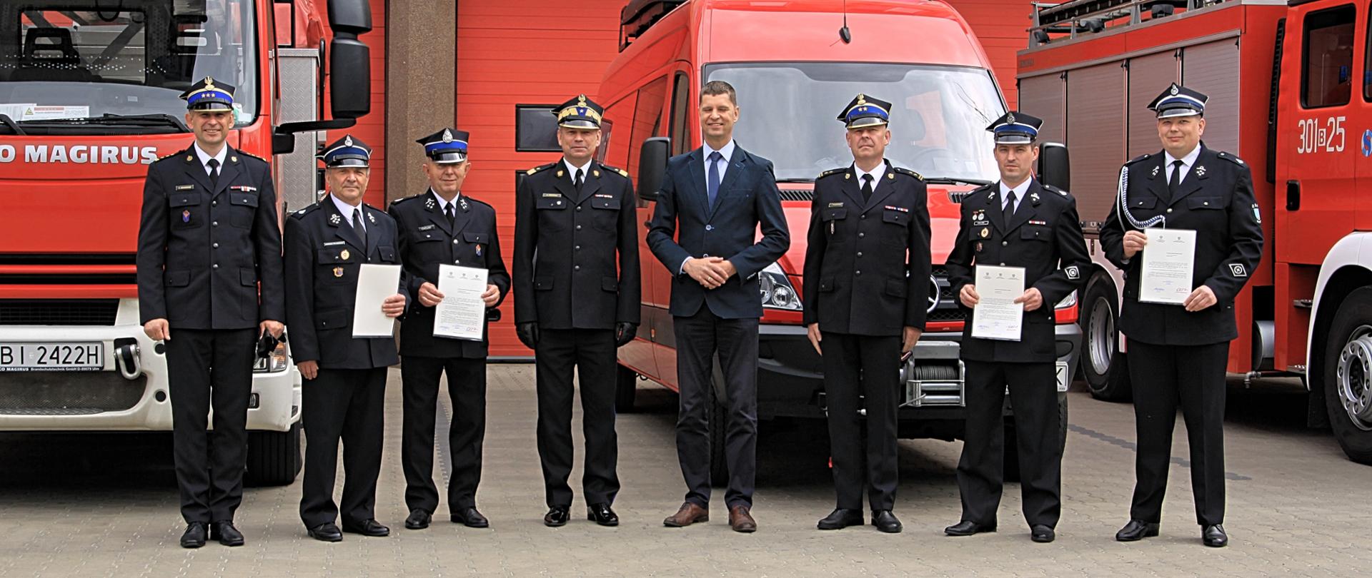 Stojący w szeregu na tle strażackich samochodów: Dariusz Piontkowski Wiceminister Edukacji i Nauki, Poseł na Sejm Rzeczypospolitej Polskiej, Zastępca Komendanta Głównego nadbrygadier Arkadiusz Przybyła, starszy brygadier Sebastian Zdanowicz Podlaski Komendant Wojewódzki Państwowej Straży Pożarnej oraz czterej druhowie z promesami.