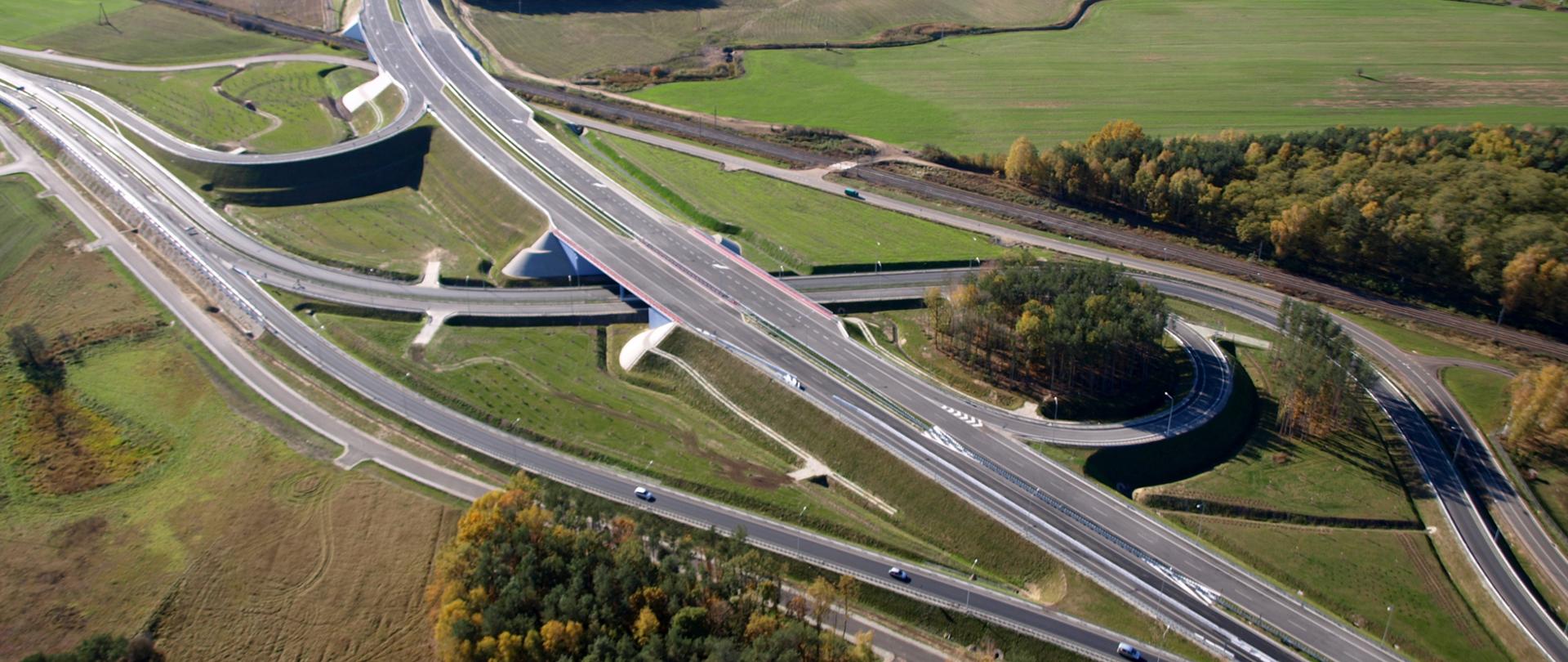 Zdjęcie przedstawiające węzeł drogowy Poznań Północ na drodze ekspresowej S11. Węzeł widoczny z powietrza przebiegający między łąkami, polami i zabudową magazynową. Na jednej z łącznic doprowadzających ruch do węzła widoczne poruszające się pojazdy.