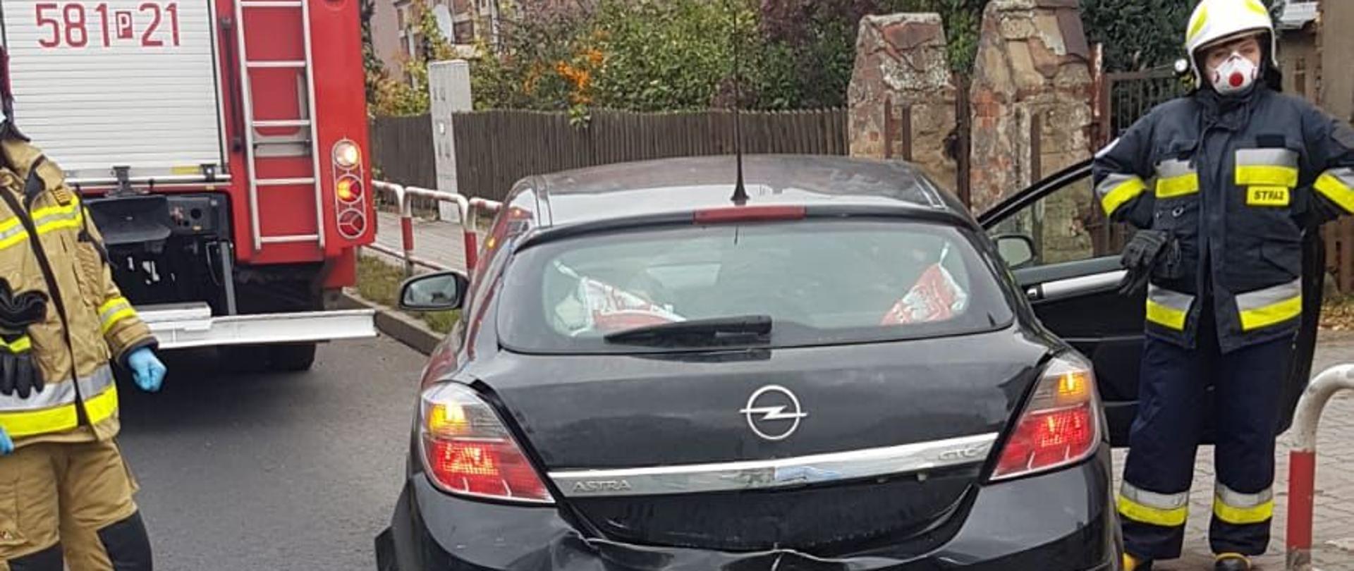 Samochód osobowy marki Opel stoi częściowo na drodze i poboczy. Widać że ma wgnieciony tył. Za nim stoi wóz strażacki a z prawej i lewej strony strażacy-ratownicy. 