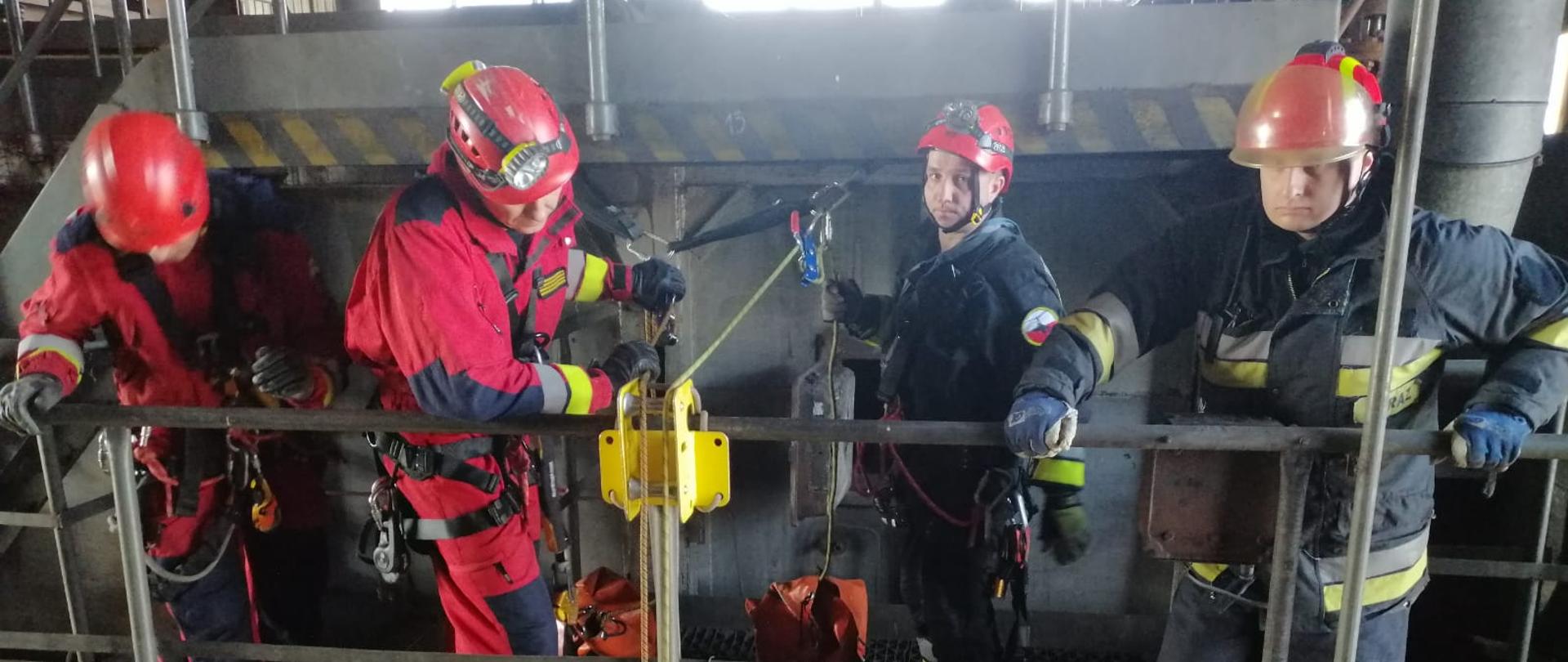 Na zdjęciu widać 4 strażaków PSP. 2 po lewej ma kombinezon wysokościowy w kolorze czerwonym oraz kaski. 2 po prawej ubrani są w ubrania specjalne w kolorze granatowym. Strażacy stoją na metalowej platformie z metalowymi barierkami. Do poręczy przymocowane jest żółte urządzenie przez, które przechodzą linki. Za strażakami widać worki na sprzęt wysokościowy. U góry zdjęcia okno. 