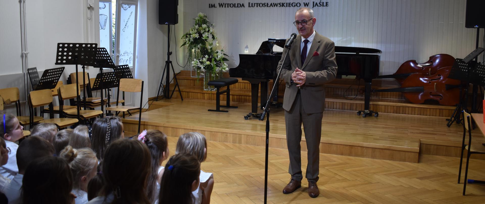 01- W auli PSM, podczas uroczystości z okazji Dnia Edukacji Narodowej, do zgromadzonej społeczności szkolnej przemawia p. Zenobiusz Kajda, wizytator Centrum Edukacji Artystycznej w Warszawie. Kolorystyka zdjęcia jest biało-brązowo-beżowo-czarna. 