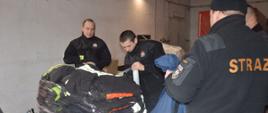 Sprzęt pożarniczy dla ukraińskich strażaków. Komenda Powiatowa PSP w Rawiczu. Strażacy przygotowują do transportu ubrania specjalne. Są one ułożone na europalecie. Jeden ze strażaków owija towar folią. W tle widoczne prądownice i węże.