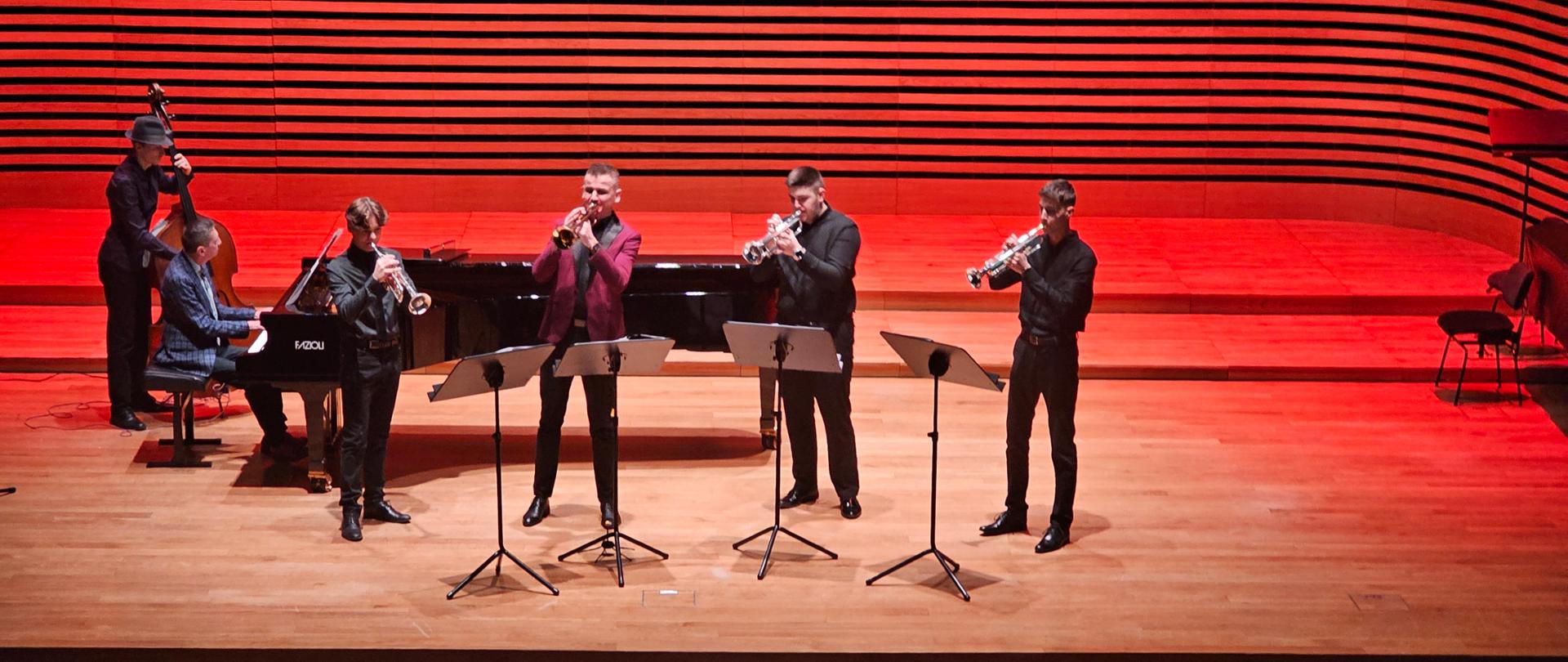 Wnętrze sali koncertowej wypełnione widzami. Na scenie pianista grający na fortepianie i kwartet dęty