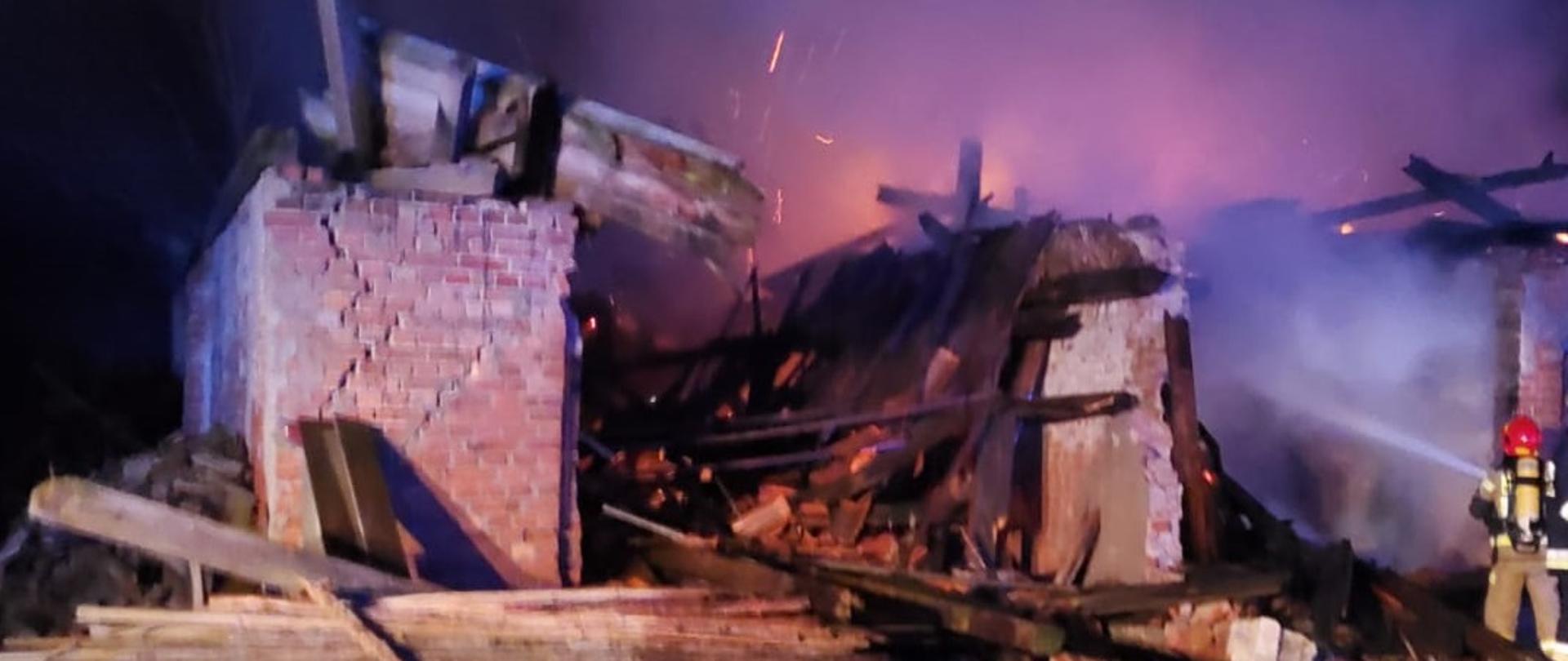 Paląca się zawalona konstrukcja budynku gospodarczego. Strażak stoi obok i podaje prądy ody na palące się elementy. Pora nocna. 