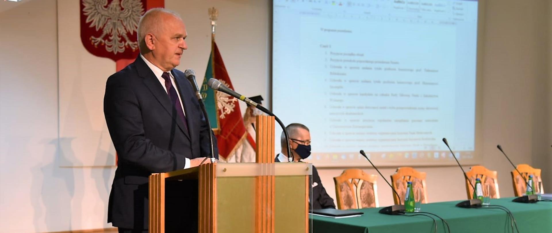 Wojewoda stoi przy mównicy z logo Uniwersytetu Zielonogórskiego. W tle wisi godło oraz stoi stół prezydialny. 