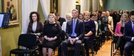 Widok z przodu na salę wypełnioną siedzącymi na ustawionych w rzędy krzesłach ludźmi, z lewej żółta ściana na której wiszą obrazy.